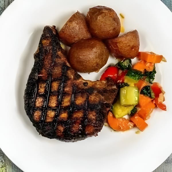 Grilled pork chop with roasted potatoes and mixed vegetables on a white plate.