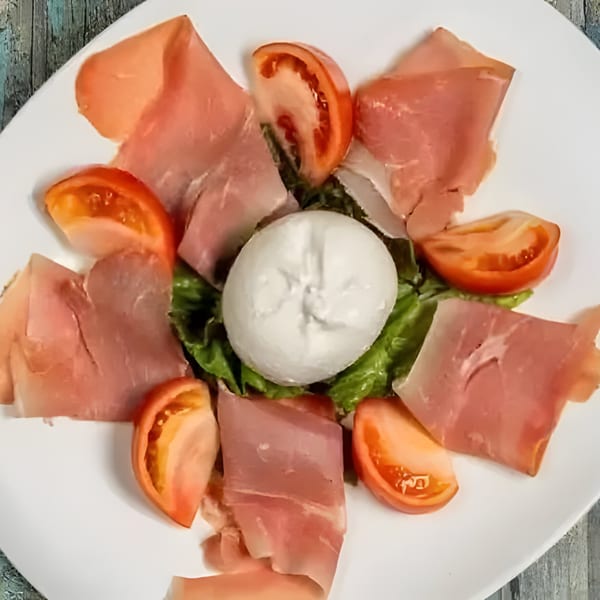 A Sage Restaurant Italian appetizer with prosciutto, burrata cheese, tomatoes, and lettuce on a white plate.
